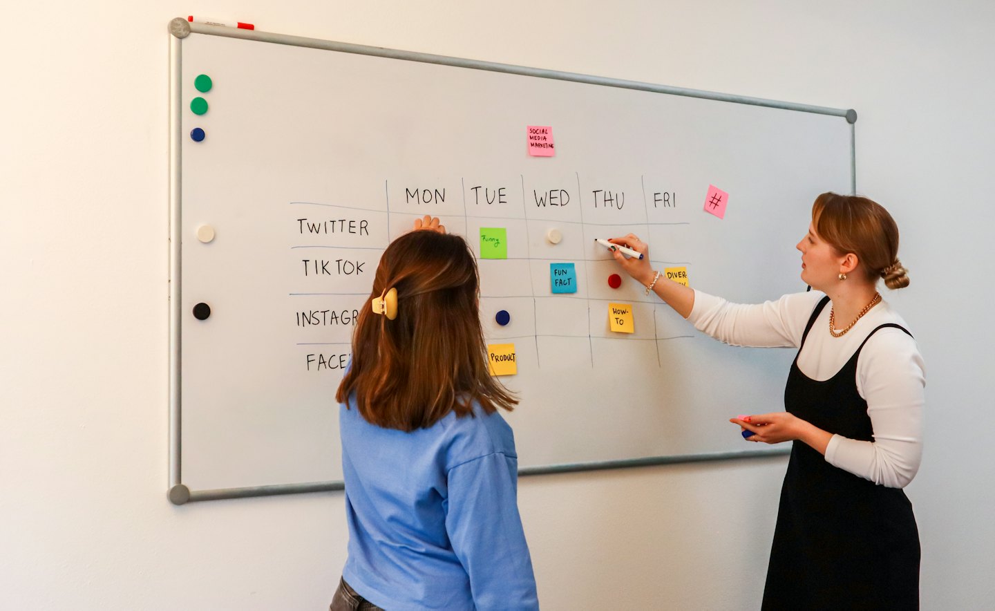 Two women planning a social media content schedule on a whiteboard.